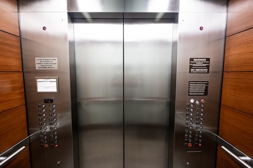 An elevator interior with sleek, metallic doors and wood panel siding. Control panels with buttons and warning signs are situated on either side, with clear labels and numbers. The lighting is bright, accentuating the polished surfaces.