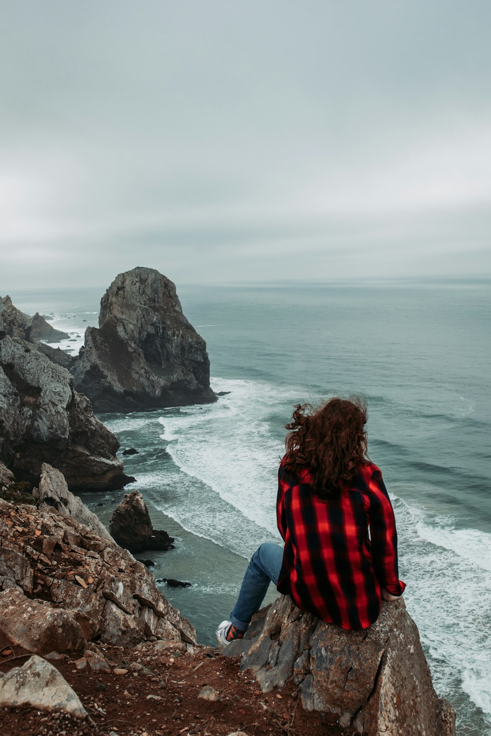 woman in red and black plaid shirt sitting on rock formation near body of water during