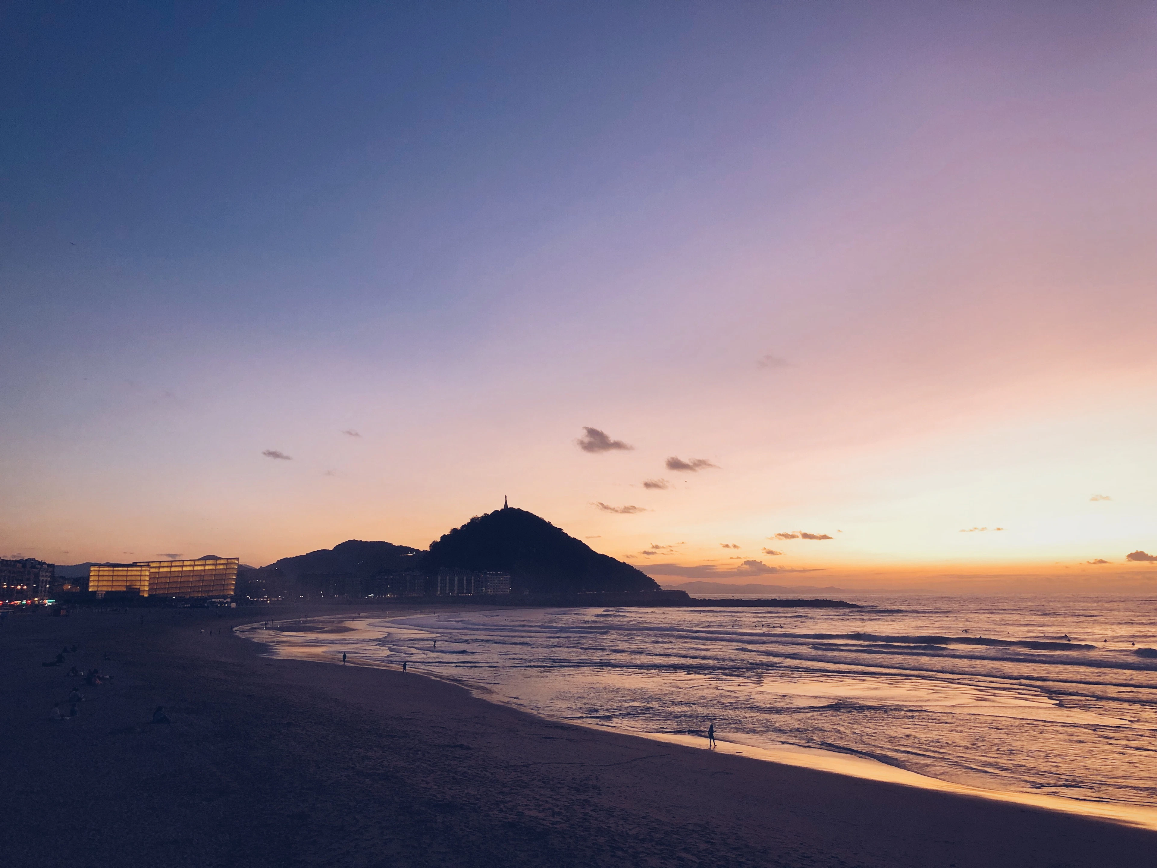 Zurriola beach in San Sebastian at sunset, wet sand catching a lilac and amber sky while the dark bulk of Monte Urgull and the lit waterfront of the Gros district stand in silhouette across the bay
