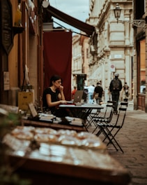 A person sits at an outdoor cafe table, using a laptop and wearing earphones. The table is set against a narrow cobblestone street lined with historic buildings. In the foreground, there is a display of pastries or baked goods, slightly out of focus. Pedestrians walk along the street, and the atmosphere suggests a casual, urban European setting.