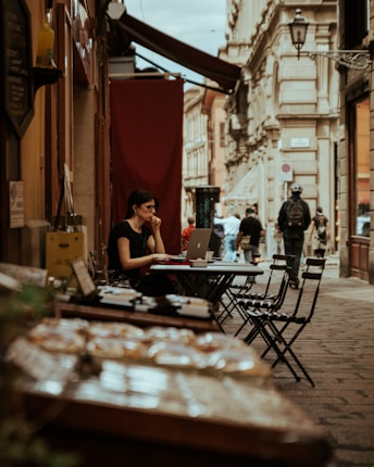 A person sits at an outdoor cafe table, using a laptop and wearing earphones. The table is set against a narrow cobblestone street lined with historic buildings. In the foreground, there is a display of pastries or baked goods, slightly out of focus. Pedestrians walk along the street, and the atmosphere suggests a casual, urban European setting.