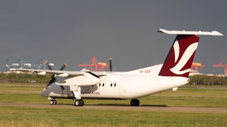 A small propeller aircraft is taxiing on a runway, with a dark sky in the background. The aircraft has a maroon and white tail fin with geometric markings. In the distance, there are industrial cranes and container terminals visible, adding a sense of scale and location.