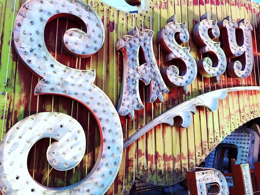 Large, ornate vintage signage displays the word 'Sassy' with illuminated bulbs on a weathered background of yellow and red vertical panels. The sign has decorative swirls and an old-fashioned, rustic feel.