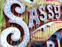 Large, ornate vintage signage displays the word 'Sassy' with illuminated bulbs on a weathered background of yellow and red vertical panels. The sign has decorative swirls and an old-fashioned, rustic feel.