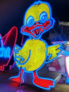 A brightly illuminated neon sign of a cartoon-style duck. The duck is outlined in vibrant blue with a yellow body and red beak. It stands in front of other neon signs, which include partially visible letters in red and blue.