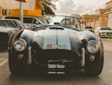 A classic sports car with a sleek black finish is parked on a sunny street surrounded by other vehicles. The design features prominent headlights and a distinctive grille. The setting suggests an upscale neighborhood with modern architecture and clear skies in the background.