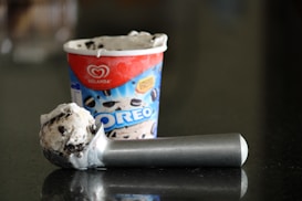 A tub of Oreo flavored ice cream with a single scoop resting on a reflective dark surface. The ice cream is creamy with visible chunks of Oreo cookies, and the scoop is metallic with a simple, sturdy design. The ice cream container features branding and a vibrant color scheme.