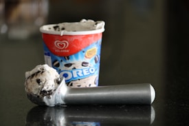 A tub of Oreo flavored ice cream with a single scoop resting on a reflective dark surface. The ice cream is creamy with visible chunks of Oreo cookies, and the scoop is metallic with a simple, sturdy design. The ice cream container features branding and a vibrant color scheme.