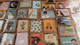 A collection of colorful greeting cards displayed on a wooden surface. The images range from illustrated faces and patterns to birthday messages with playful and humorous designs. Some cards feature people in various poses, while others have drawings like a cactus, a giraffe, and modern art-style illustrations.