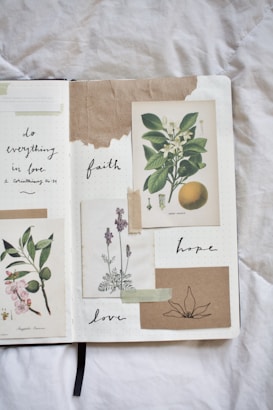 A journal spread features botanical illustrations and handwritten words. The left side displays a drawing of flowers and leaves, while the right side contains images of a sweet orange and lavender. Words like 'faith,' 'hope,' and 'love' are thoughtfully written around the illustrations. The page has a textured and artistic feel with the use of tape and torn paper.