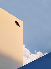 A minimalist architectural scene with a beige building featuring a singular circular opening, set against a backdrop of a bright blue sky with fluffy clouds. The lower right corner shows a geometric blue structure, creating a contrast with the soft clouds.