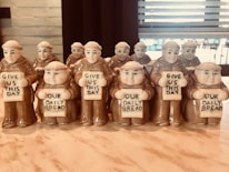A collection of small monk figurines holding plaques with the words 'Give Us This Day' and 'Our Daily Bread'. The figurines are neatly arranged in rows on a marble countertop with a blurred background of window blinds.