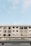 A modern, multi-story building with a symmetrical facade featuring numerous windows. The structure is primarily white with black window frames. The lower portion of the image shows a sign with the word 'CARPENTER'. The sky above is clear, with soft cloud formations, conveying a calm atmosphere.