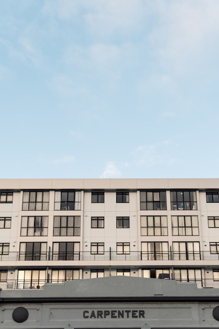 A modern, multi-story building with a symmetrical facade featuring numerous windows. The structure is primarily white with black window frames. The lower portion of the image shows a sign with the word 'CARPENTER'. The sky above is clear, with soft cloud formations, conveying a calm atmosphere.
