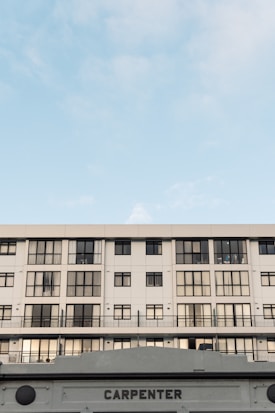 A modern, multi-story building with a symmetrical facade featuring numerous windows. The structure is primarily white with black window frames. The lower portion of the image shows a sign with the word 'CARPENTER'. The sky above is clear, with soft cloud formations, conveying a calm atmosphere.