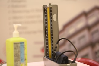 A sphygmomanometer and a pump bottle are placed on a light-colored surface. The sphygmomanometer has a gauge with clearly marked increments and a rubber tube attached to it. The background features a blurry image of a building.