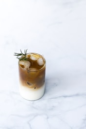 A glass of iced coffee with a creamy milk layer at the bottom, topped with a brown coffee layer. Ice cubes float at the top, and a sprig of rosemary adds a decorative touch. The background is a light marble surface.