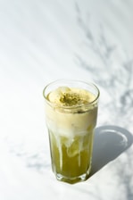 A tall glass filled with a layered beverage, featuring a frothy cream on top sprinkled with green powder, possibly matcha. The bottom layer is a vibrant green liquid. Soft shadows are subtly cast on the white surface, suggesting natural lighting.