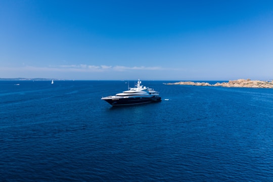 A large luxury yacht is floating in a vast expanse of clear blue ocean, with smaller boats visible in the distance. The sky is bright blue and cloudless, meeting the horizon where the sea meets a distant landmass with rocky shores.