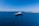 A large luxury yacht is floating in a vast expanse of clear blue ocean, with smaller boats visible in the distance. The sky is bright blue and cloudless, meeting the horizon where the sea meets a distant landmass with rocky shores.