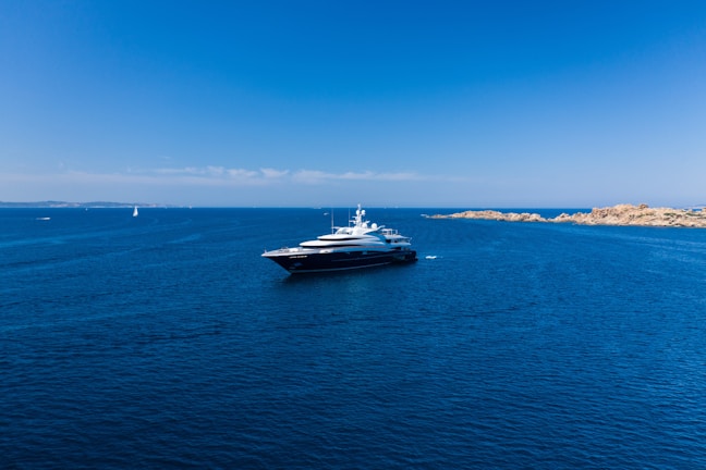A large luxury yacht is floating in a vast expanse of clear blue ocean, with smaller boats visible in the distance. The sky is bright blue and cloudless, meeting the horizon where the sea meets a distant landmass with rocky shores.