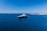 A large luxury yacht is floating in a vast expanse of clear blue ocean, with smaller boats visible in the distance. The sky is bright blue and cloudless, meeting the horizon where the sea meets a distant landmass with rocky shores.