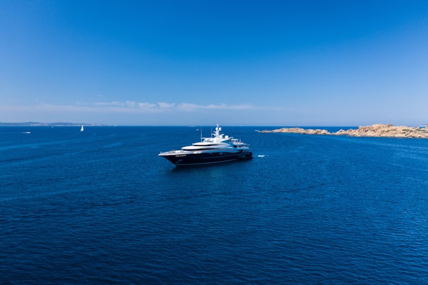 A large luxury yacht is floating in a vast expanse of clear blue ocean, with smaller boats visible in the distance. The sky is bright blue and cloudless, meeting the horizon where the sea meets a distant landmass with rocky shores.