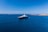 A large luxury yacht is floating in a vast expanse of clear blue ocean, with smaller boats visible in the distance. The sky is bright blue and cloudless, meeting the horizon where the sea meets a distant landmass with rocky shores.