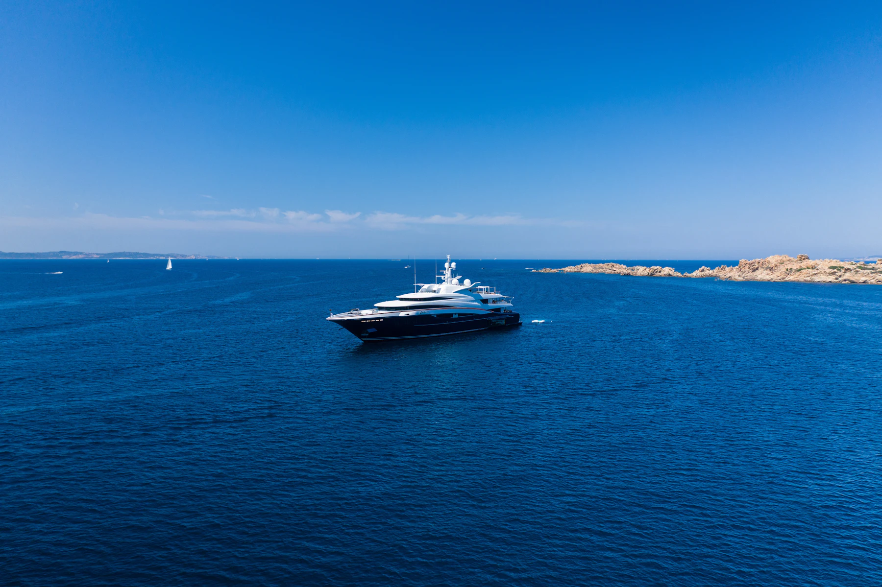 A yacht running along a rocky coastline in open blue water.