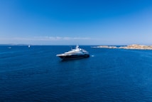 A large luxury yacht is floating in a vast expanse of clear blue ocean, with smaller boats visible in the distance. The sky is bright blue and cloudless, meeting the horizon where the sea meets a distant landmass with rocky shores.