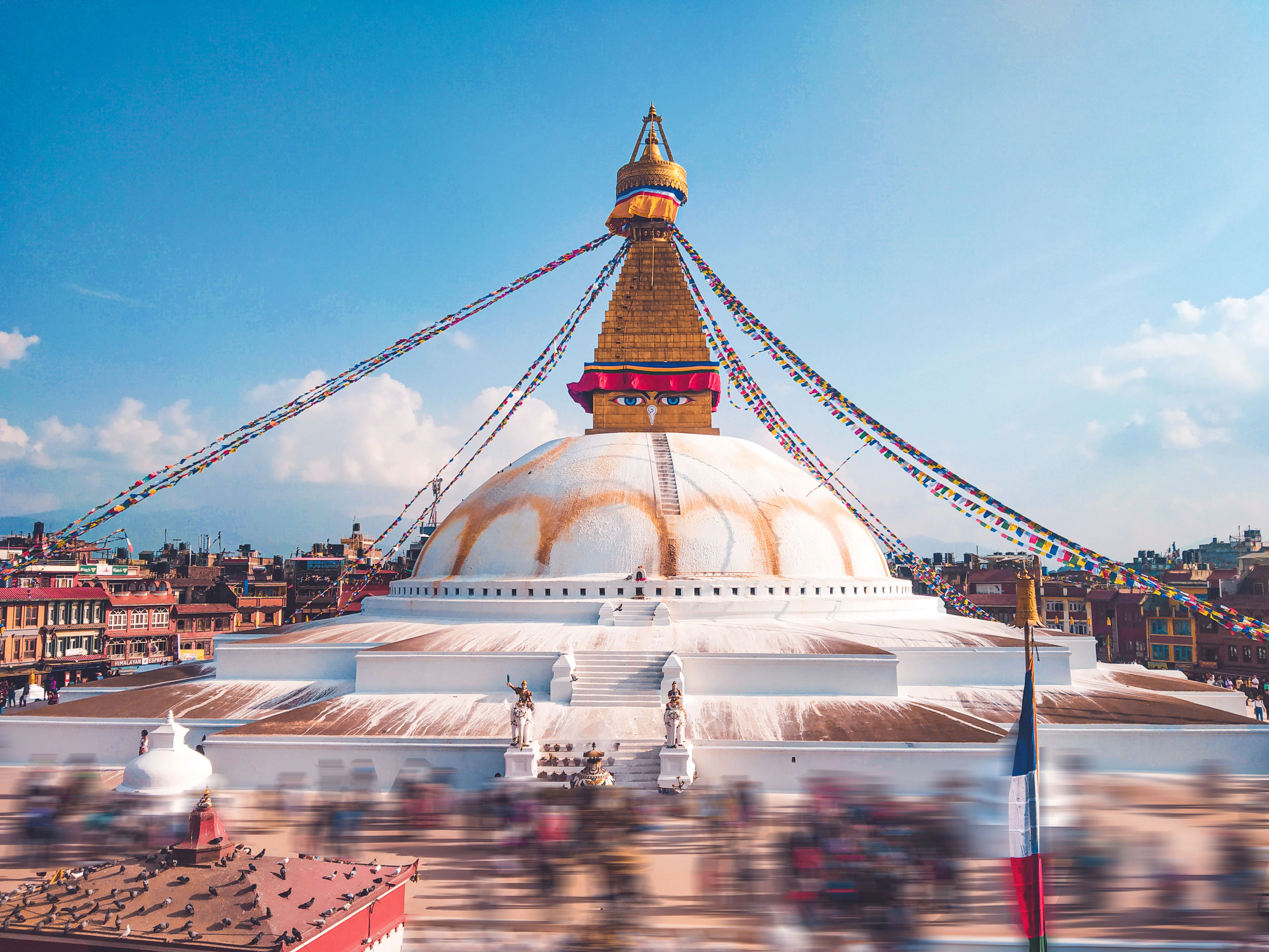 The gold-spired dome of Boudhanath Stupa rises above prayer flags and rooftops under a clear blue sky.