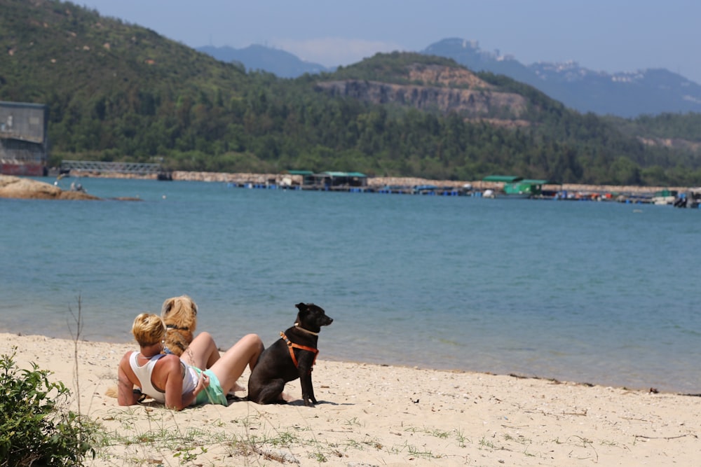 woman in black tank top lying on white sand beside black short coat dog during daytime