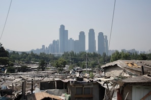 A contrast between an urban slum in the foreground with makeshift houses and satellite dishes, and a distant skyline of modern skyscrapers under a hazy sky. Power lines cross the scene, with trees scattered among the buildings.