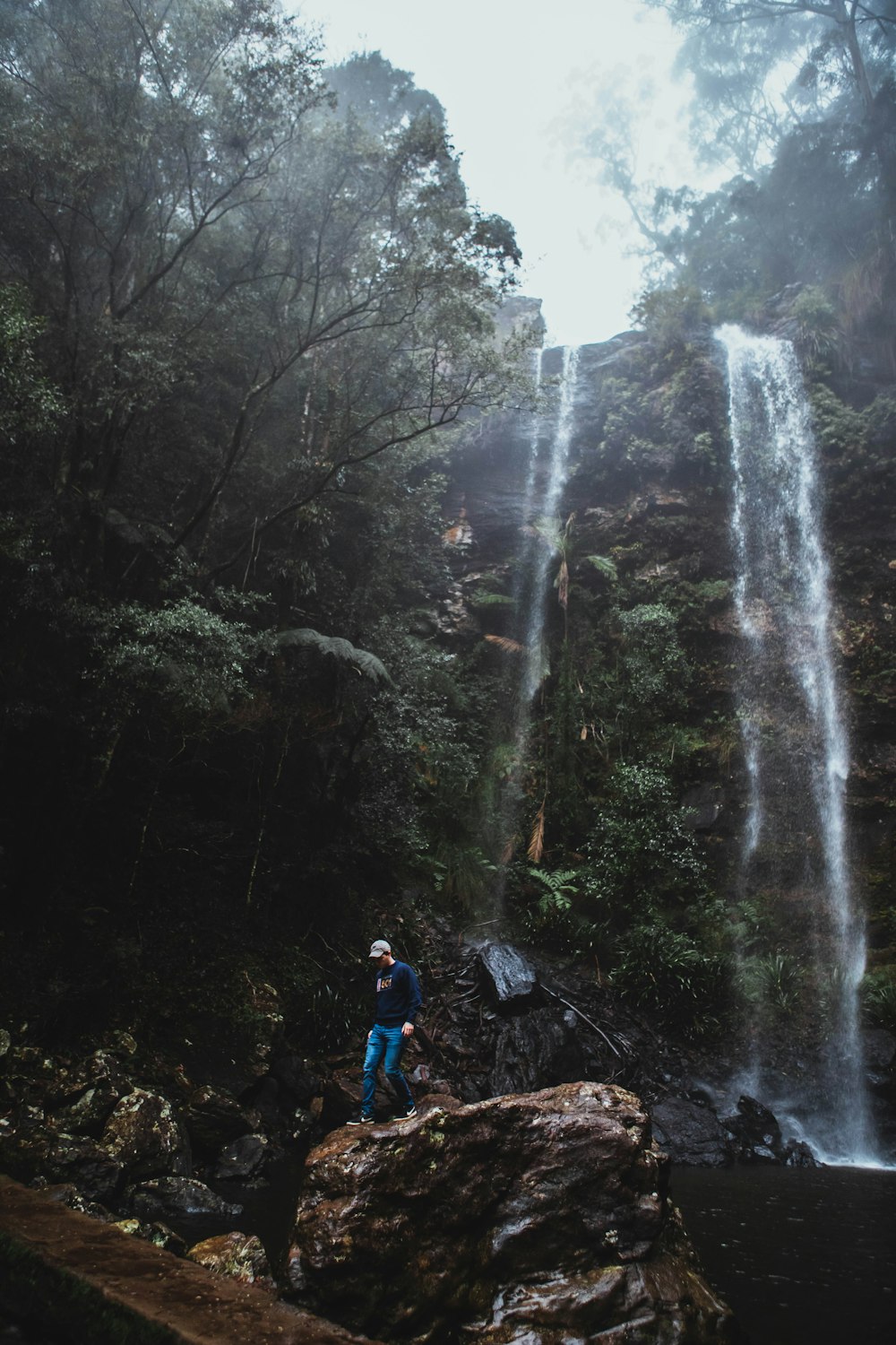 man in blue jacket and blue denim jeans standing on brown rock near waterfalls during daytime