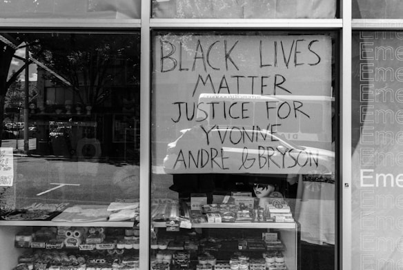 A shop window features a prominent handwritten sign stating 'Black Lives Matter Justice for Yvonne, Andre & Bryson'. The window displays various merchandise, including snacks and small toys. Reflections on the glass showcase street details and nearby buildings.