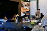 A group of people are sitting around a conference table in a meeting room. A woman is actively speaking while holding a laptop. In the background, there is a banner for a 'UX Training Series' and whiteboards covered with diagrams and notes. Several objects are on the table, including papers, notebooks, electronic devices, and drinks.
