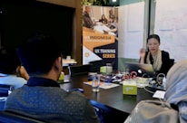 A group of people are sitting around a conference table in a meeting room. A woman is actively speaking while holding a laptop. In the background, there is a banner for a 'UX Training Series' and whiteboards covered with diagrams and notes. Several objects are on the table, including papers, notebooks, electronic devices, and drinks.