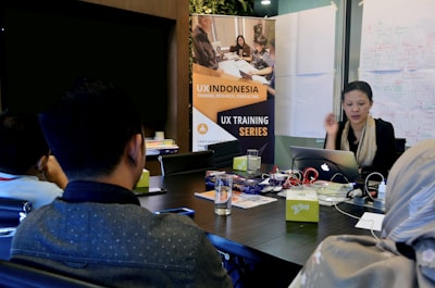 A group of people are sitting around a conference table in a meeting room. A woman is actively speaking while holding a laptop. In the background, there is a banner for a 'UX Training Series' and whiteboards covered with diagrams and notes. Several objects are on the table, including papers, notebooks, electronic devices, and drinks.