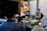 A group of people are sitting around a conference table in a meeting room. A woman is actively speaking while holding a laptop. In the background, there is a banner for a 'UX Training Series' and whiteboards covered with diagrams and notes. Several objects are on the table, including papers, notebooks, electronic devices, and drinks.