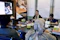A group of people are gathered in a conference room, engaged in a UX training session. Two women are seated at a table with laptops, participating in the training. A large poster on the wall advertises UX Indonesia and a UX training series. Papers with diagrams are taped to the wall, and various electronic devices and cups are on the table.