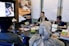 A group of people are gathered in a conference room, engaged in a UX training session. Two women are seated at a table with laptops, participating in the training. A large poster on the wall advertises UX Indonesia and a UX training series. Papers with diagrams are taped to the wall, and various electronic devices and cups are on the table.