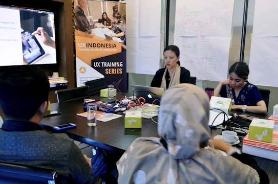 A group of people are gathered in a conference room, engaged in a UX training session. Two women are seated at a table with laptops, participating in the training. A large poster on the wall advertises UX Indonesia and a UX training series. Papers with diagrams are taped to the wall, and various electronic devices and cups are on the table.