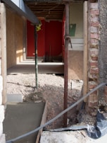 A partially constructed or renovated room with exposed brickwork and wooden paneling. The floor has been removed, revealing an uneven dirt surface. Temporary wooden and metal supports are placed at various points. Debris is scattered around the area, and a bright red wall is visible at the back of the room.