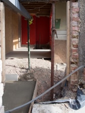 A partially constructed or renovated room with exposed brickwork and wooden paneling. The floor has been removed, revealing an uneven dirt surface. Temporary wooden and metal supports are placed at various points. Debris is scattered around the area, and a bright red wall is visible at the back of the room.