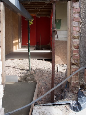 A partially constructed or renovated room with exposed brickwork and wooden paneling. The floor has been removed, revealing an uneven dirt surface. Temporary wooden and metal supports are placed at various points. Debris is scattered around the area, and a bright red wall is visible at the back of the room.