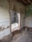 A partially demolished room with exposed brick walls and stripped tiles. Electrical conduits are visible, indicating ongoing construction or renovation. The interior view reveals debris, wires, and unfinished surfaces. A wall opening leads to another space that appears incomplete.