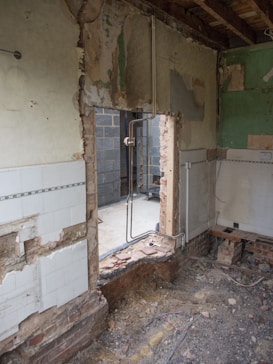 A partially demolished room with exposed brick walls and stripped tiles. Electrical conduits are visible, indicating ongoing construction or renovation. The interior view reveals debris, wires, and unfinished surfaces. A wall opening leads to another space that appears incomplete.