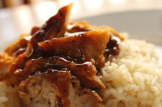 A close-up of a dish featuring cooked rice and fried chicken pieces topped with a glossy brown sauce. The chicken appears crispy with visible texture, and the rice grains are fluffy and well-cooked.