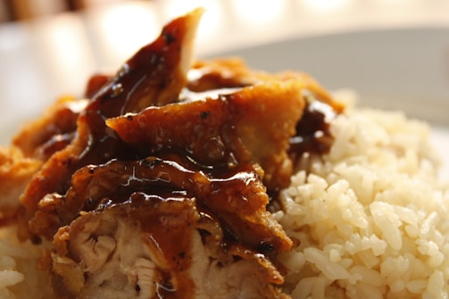 A close-up of a dish featuring cooked rice and fried chicken pieces topped with a glossy brown sauce. The chicken appears crispy with visible texture, and the rice grains are fluffy and well-cooked.