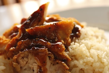 A close-up of a dish featuring cooked rice and fried chicken pieces topped with a glossy brown sauce. The chicken appears crispy with visible texture, and the rice grains are fluffy and well-cooked.